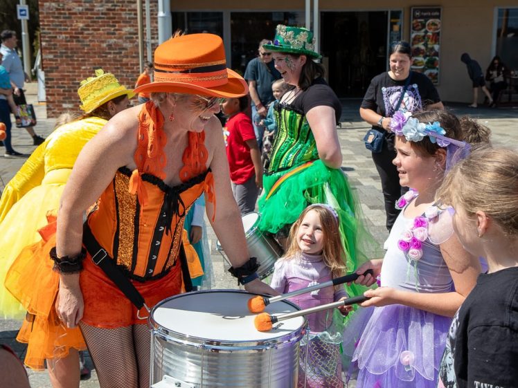 Image of A woman in an orange costume plays a drum outdoors while smiling at children dressed in colourful costumes, including fairy and princess outfits. Other adults and kids in festive clothes watch and enjoy the event.