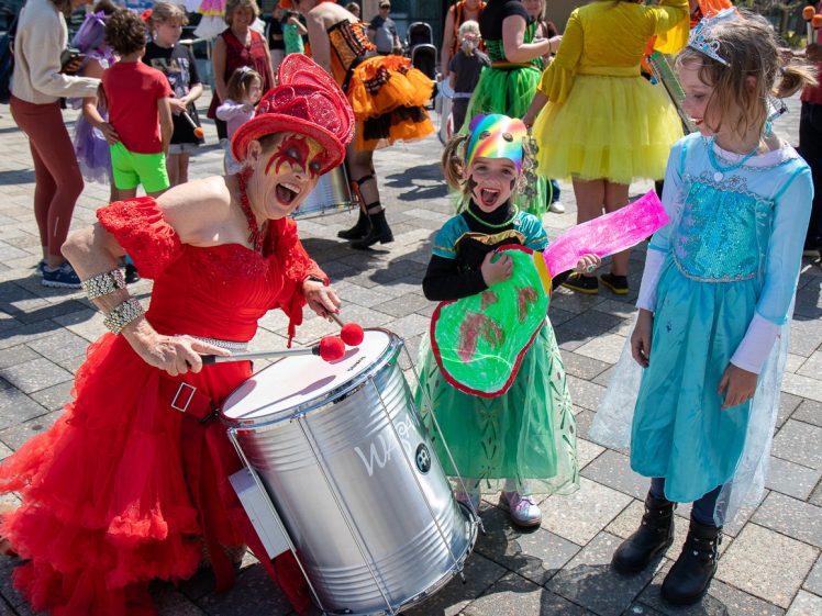 Image of A woman in a red costume plays a drum next to two smiling children in colourful costumes at an outdoor event with other people in fancy dress in the background.
