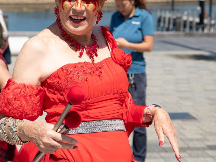 Image of A person in an elaborate red costume and face paint smiles whilst playing a drum outdoors. Other people and water are visible in the background on a sunny day.