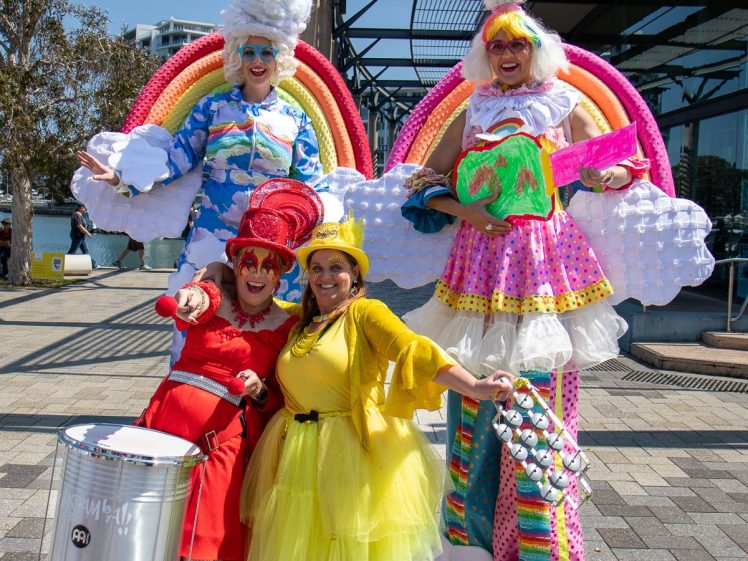 Image of Four people in colourful, whimsical costumes pose cheerfully outdoors. Two wear tall rainbow and cloud outfits on stilts, while the other two wear bright red and yellow costumes and smile energetically at the camera.