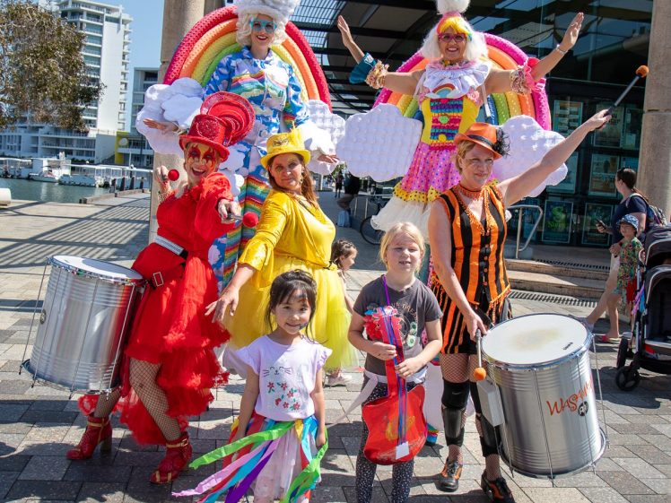 Image of A group of performers in colourful costumes, including two on stilts dressed as rainbows and clouds, pose with children holding ribbons and shakers outside a building on a sunny day. Two people play large drums.