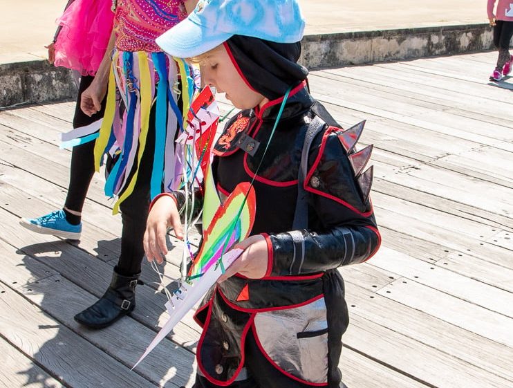 Image of A child in a dragon costume and cap walks on a wooden walkway, holding colourful paper. An adult in a vibrant, rainbow outfit and crown smiles nearby. Other people are visible in the background.