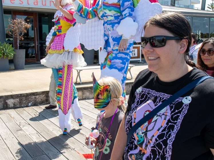 Image of Two colourful performers on stilts, dressed as rainbows and clouds, entertain a crowd on a promenade. A smiling woman and a child in a rainbow mask and outfit walk in the foreground. A block of flats is in the background.