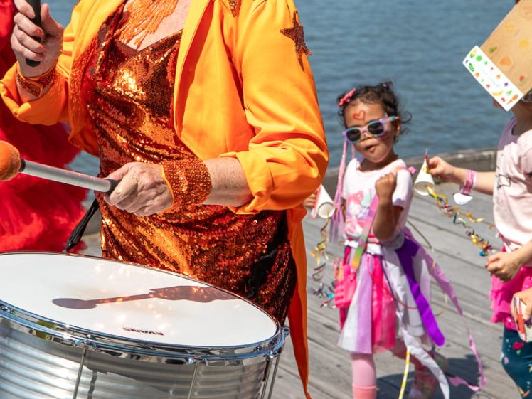Image of A smiling drummer in a bright orange carnival costume leads a festive parade near the water, followed by children in colourful outfits and face paint.