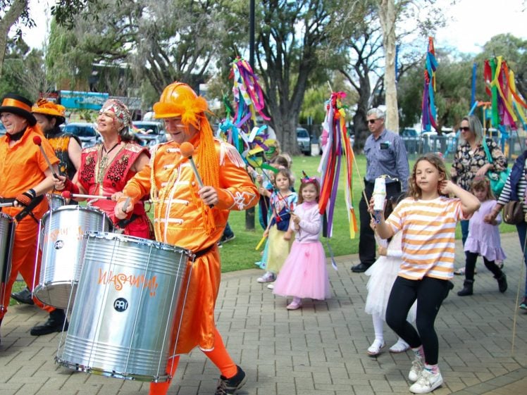 Image of A lively outdoor parade features people in bright costumes playing drums and maracas, children in fairy outfits, and others waving colourful ribbons, all walking together on a park pathway lined with trees.