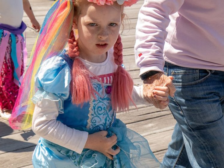 Image of A young girl in a blue princess fancy dress with pink plaited hair and a flower crown holds hands with an adult on a sunny wooden walkway. Another child in fancy dress and a pushchair are in the background.