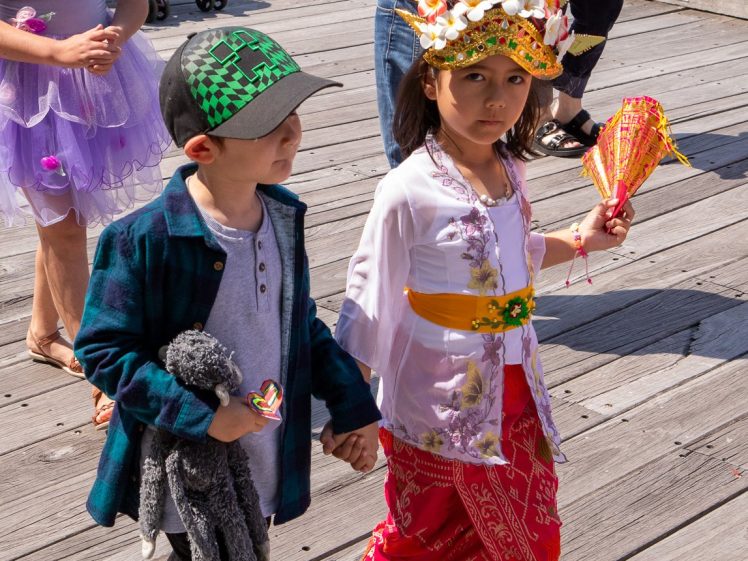Image of Two children walk hand in hand on a wooden jetty. One wears a cap, checked shirt, and holds a soft toy, while the other wears a vibrant traditional outfit with a decorated headdress and holds colourful fans.
