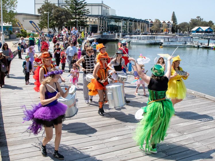 Image of A colourful parade of people in vibrant costumes play drums and other instruments whilst walking along a wooden boardwalk beside a marina with boats on a sunny day. Spectators follow and watch the lively procession.
