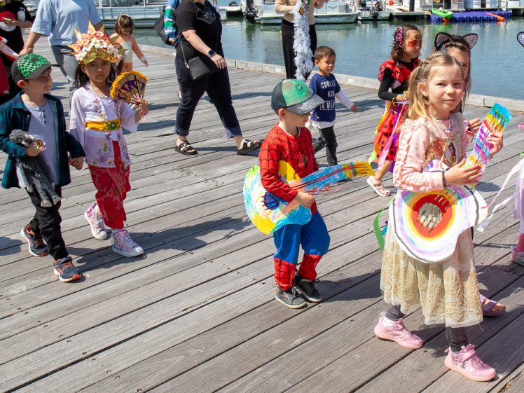 Image of A group of young children in colourful costumes walk along a wooden quay, each holding a cardboard guitar decorated with bright colours. Boats and water are visible in the background on a sunny day.