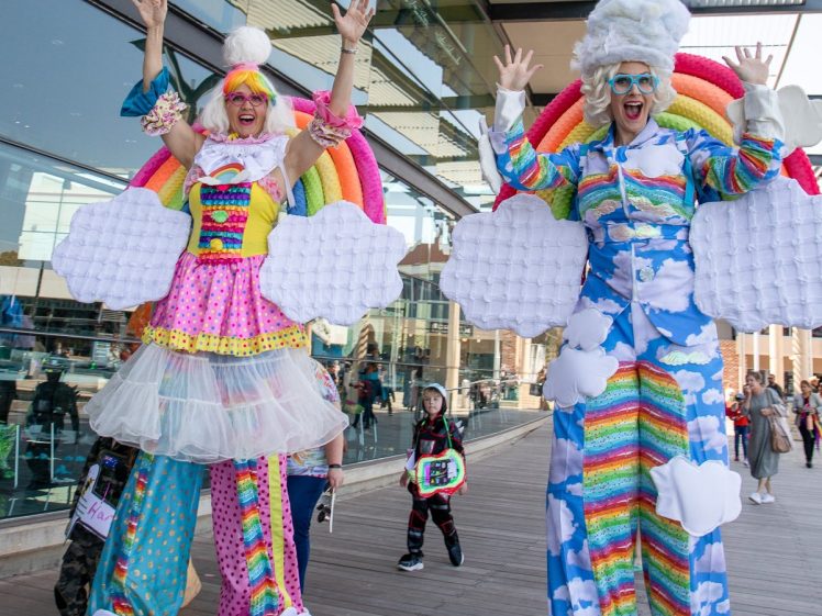 Image of Two people on stilts dressed in colourful, cloud and rainbow-themed costumes pose with raised arms outside a modern glass building. A child in fancy dress walks behind them on a wooden walkway.