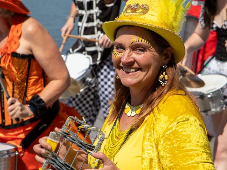 Image of A woman dressed in bright yellow, smiling and playing a handheld percussion instrument at an outdoor waterfront event, with other costumed musicians in colourful outfits behind her.