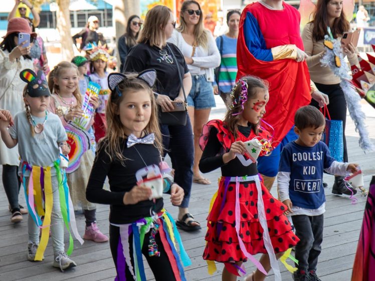 Image of Children in colourful costumes, including a cat and ladybird outfit, walk in a parade. Adults, some in fancy dress, watch and take photos. The atmosphere is festive and lively, with decorations and smiling faces.