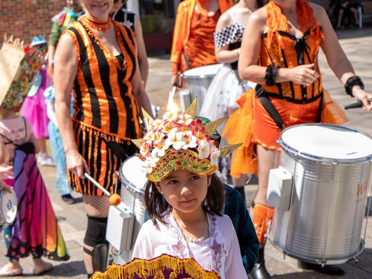 Image of A young girl in a decorative hat and white dress holds a fan whilst standing in front of a group of smiling adults dressed in bright orange and black costumes, playing drums outdoors on a sunny day.