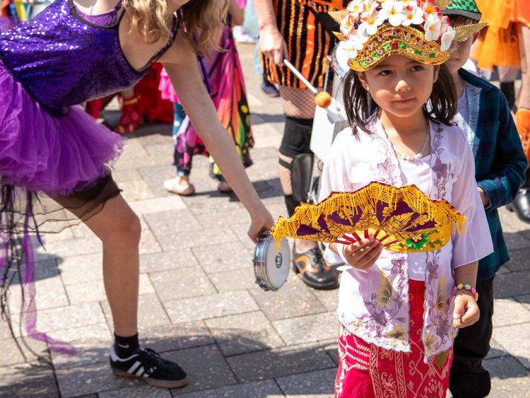 Image of A young girl in colourful traditional attire holds a decorated fan, while behind her, a smiling woman in a purple tutu and flower crown leans towards the camera, surrounded by people in bright costumes at an outdoor event.