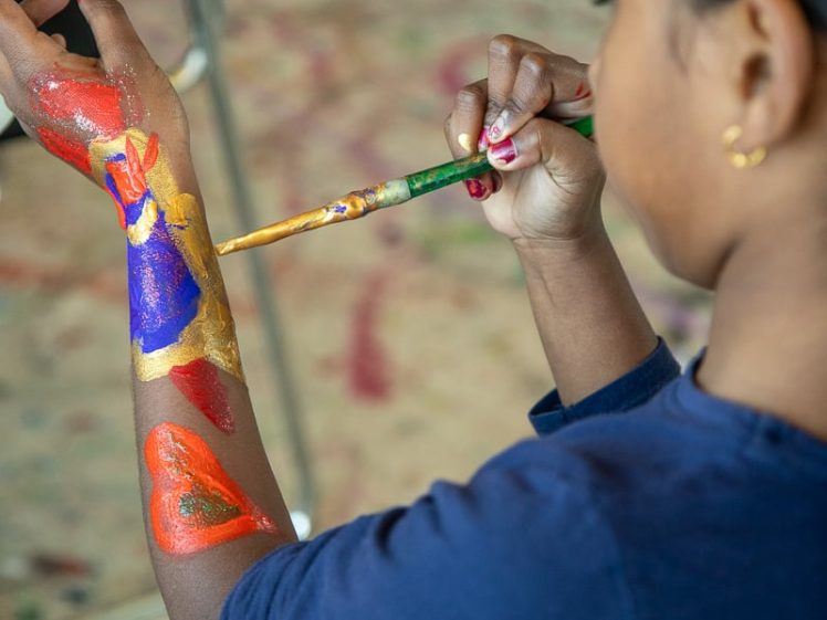 Image of A child wearing a navy blue shirt paints colourful designs on their forearm with a brush, including red and blue shapes, whilst sitting indoors.