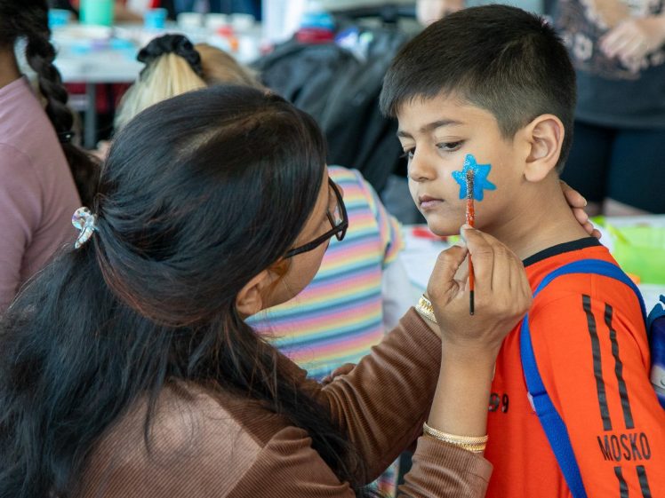 Image of A woman paints a blue star on a boy’s cheek at an indoor event. The boy is wearing an orange shirt and looks to the side whilst the woman, holding a brush, carefully adds details to the star.