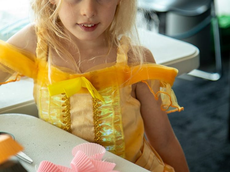 Image of A young girl in a yellow princess dress and tiara sits at a table with pink cake cases, striped straws, and party supplies, looking at the camera.