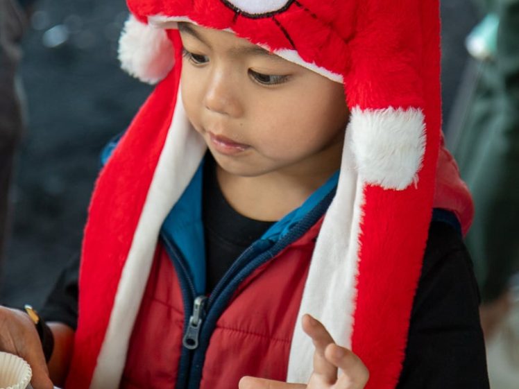 Image of A young child wearing a red Spider-Man hat and red waistcoat sits at a table, looking at empty white paper cake cases in front of him.