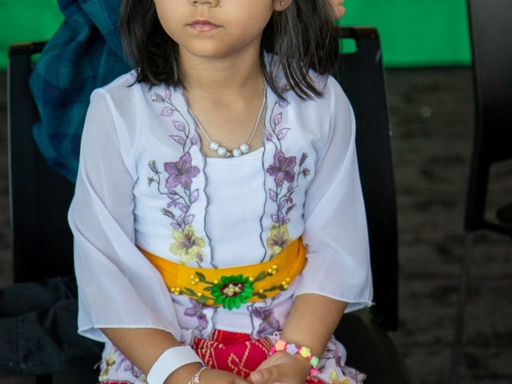Image of A young girl wearing a traditional Balinese outfit and a decorative gold headdress with white and red flowers sits on a chair, looking thoughtful. A green banner is visible in the background.