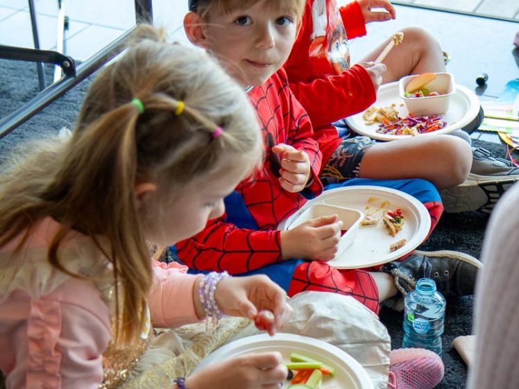 Image of Three children in fancy dress sit on the floor eating food from paper plates. One wears a pink and gold dress, another a Spider-Man costume and green cap, and the third a red superhero outfit. Bottled water is beside them.
