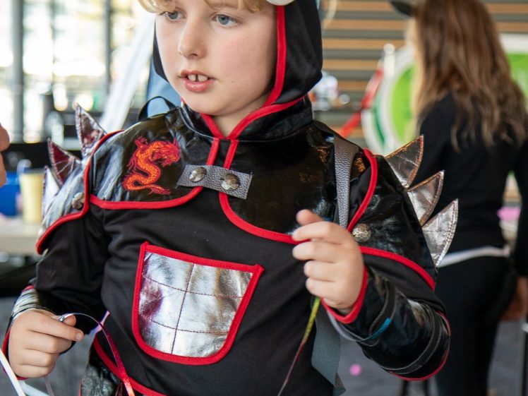 Image of A young child in a black and red knight costume with silver armour details and a light blue hat stands indoors, holding craft supplies at a busy table.