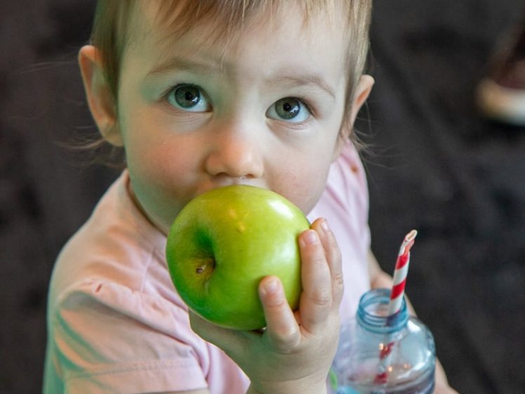Image of A young child with light brown hair holds a green apple to their mouth and a water bottle with a striped straw. The child is wearing a pale pink shirt and looking up at the camera.