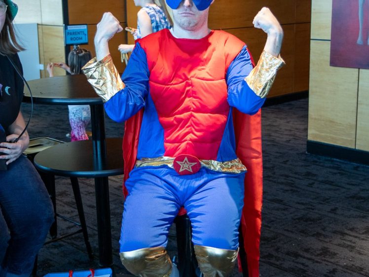 Image of A man in a blue and red superhero costume with a cape, gold cuffs, and a blue mask sits on a stool, flexing his arms and making a serious face. A Thomas the Tank Engine gift bag is on the floor beside him.