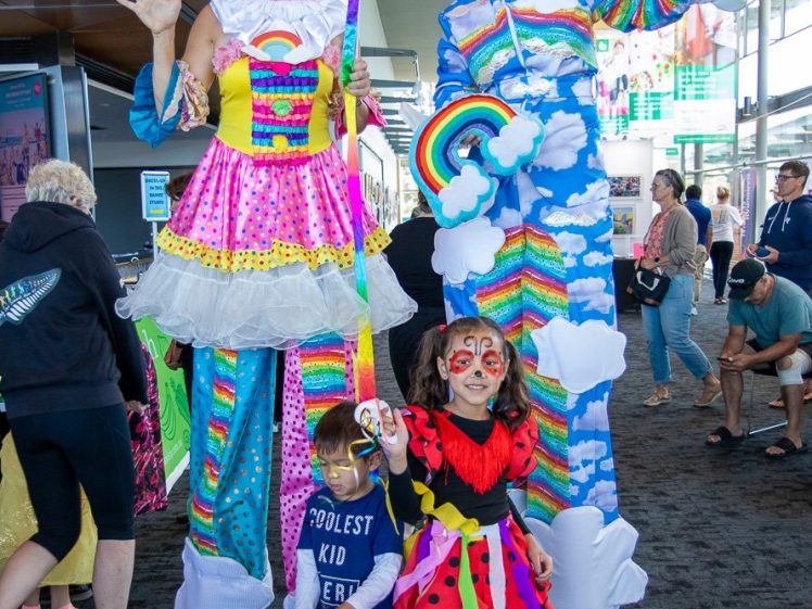 Image of Two adults on stilts in colourful, rainbow-themed costumes with clouds and rainbows stand indoors. Two children, one with a rainbow painted face and the other in a ladybird costume, pose and smile in front of them. People walk in the background.