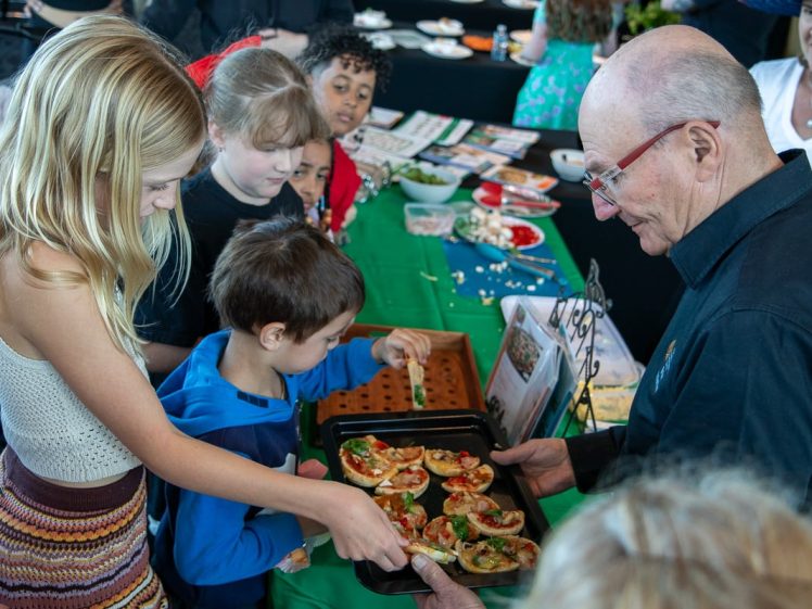 Image of A man holds a tray of mini pizzas as children eagerly take pieces at a crowded event table covered with food and crafts. Adults and children watch, and the atmosphere is lively and festive.
