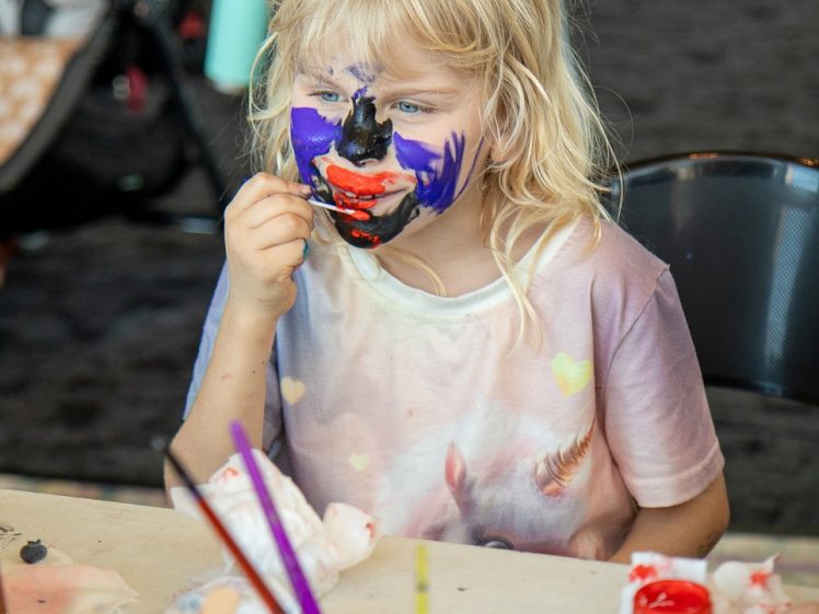 Image of A young child with long blonde hair and colourful face paint sits at a table with art supplies and paint jars, holding a paintbrush to their face in a lively indoor setting.
