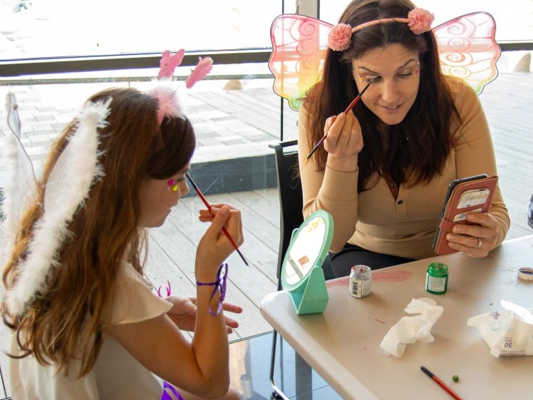 Image of A woman and a girl in fairy costumes sit at a table, painting their faces with brushes. The woman uses her mobile as a mirror, while the girl paints her cheek. Art supplies and a small mirror are on the table.