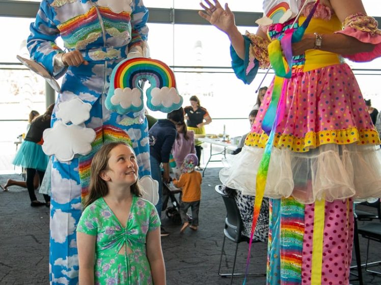Image of Two performers on stilts dressed in bright rainbow and cloud costumes smile and interact with a young girl in a green dress at an indoor event with tables of food.