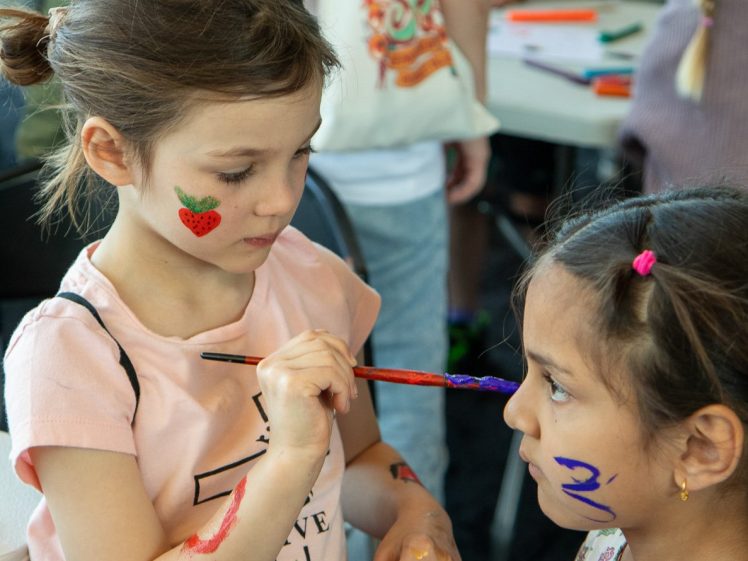 Image of Two young girls are face painting; one girl gently paints on the other’s face with a brush. Both have colourful designs on their faces and hands. Children and art supplies are visible in the background.