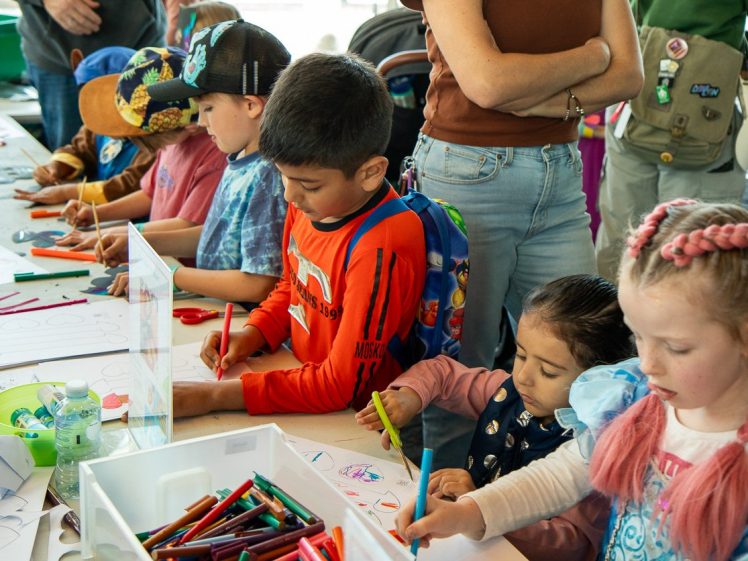 Image of Children sit and stand around a table, colouring with felt-tip pens and crayons on sheets of paper. An adult stands nearby, supervising. Art supplies are scattered on the table. The scene is lively and creative.