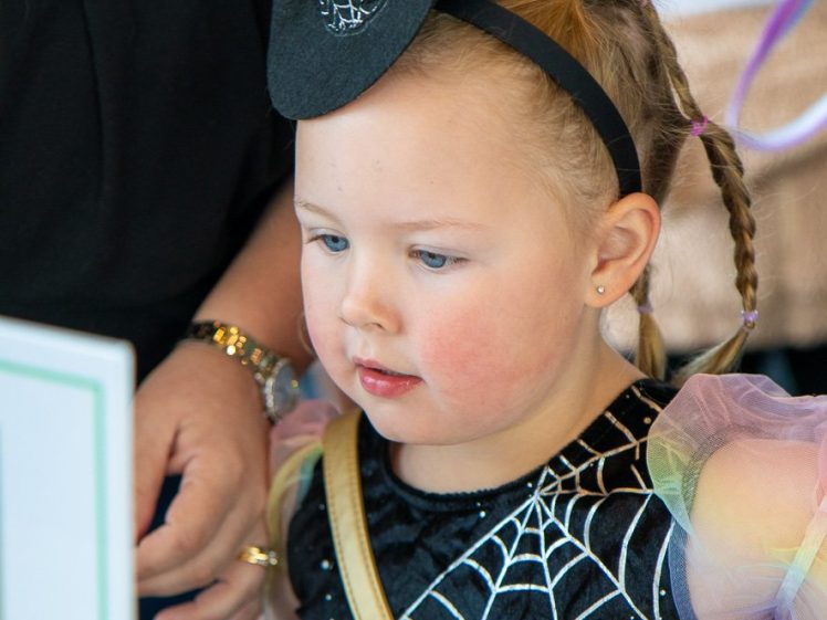 Image of A young child dressed in a black costume with spiderweb designs and a small witch hat headband looks intently at something off-camera, with an adult standing close by.