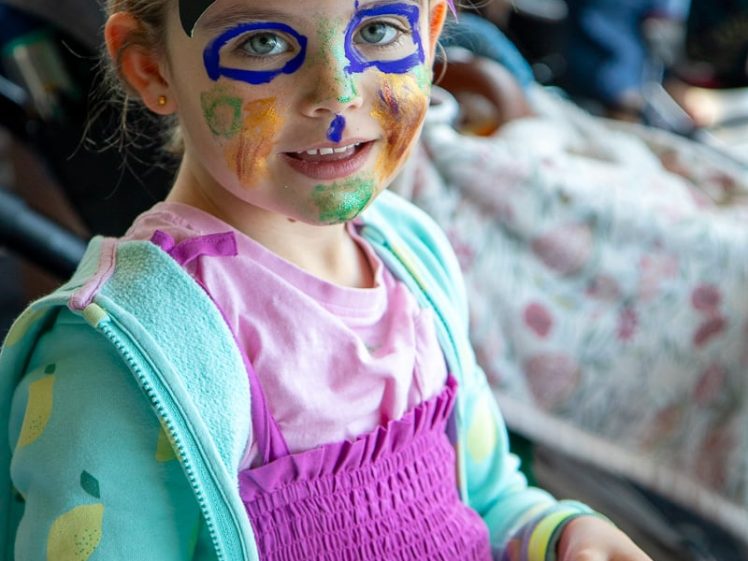Image of A young child with colourful face paint wears a handmade, multicoloured paper hat. She smiles at the camera whilst sitting at a table, holding paper craft materials. She is dressed in a blue jacket with a lemon pattern and a pink top.
