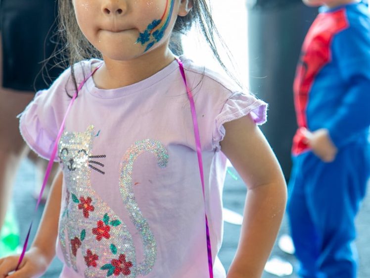 Image of A young girl with rainbow face paint wears a purple shirt with a glittery cat design and holds a red shiny object. In the background, a child dressed in a superhero costume with face paint is visible.