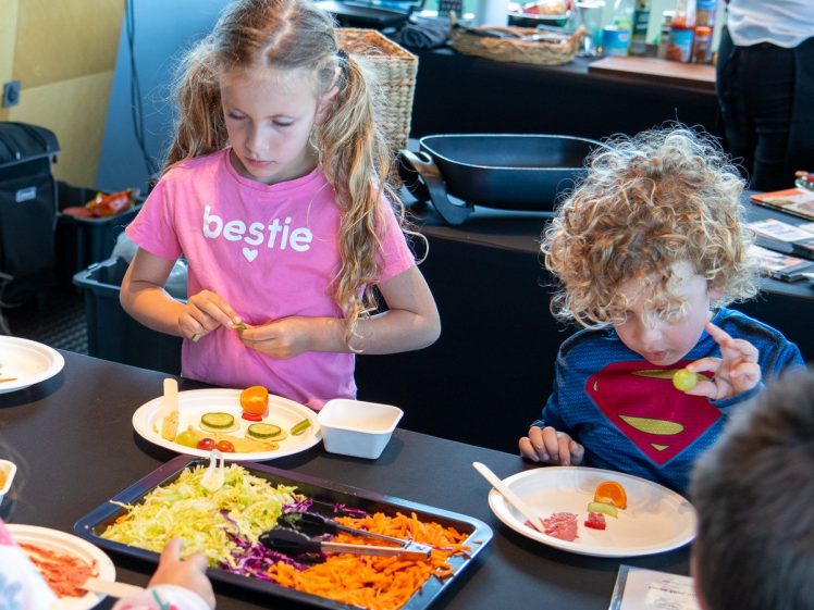 Image of Two young children prepare food at a table, arranging sliced vegetables on plates. One girl in a pink bestie shirt concentrates on her plate, while a boy in a Superman outfit examines a cucumber slice.