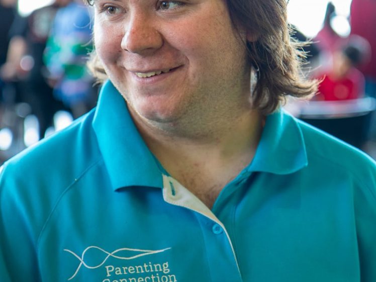 Image of A person wearing a gold glittery hat and a blue Parenting Connection polo shirt smiles whilst standing indoors at a social event.