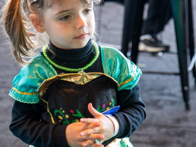 Image of Young girl with light brown hair in plaits wearing a jewelled tiara and a green princess dress, standing indoors with her hands clasped, looking slightly to the side.