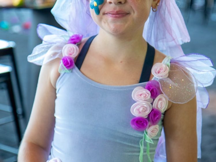 Image of A smiling child wearing a pastel fairy costume with flower decorations, a flower crown, and a veil, along with a painted blue flower on their cheek, poses indoors at what appears to be a festive event.