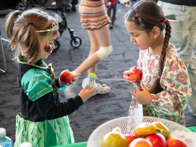 Image of Two young girls stand facing each other, each holding a red apple. One girl is dressed in a costume with a crown, and the other wears a floral dress. A bowl of fruit sits on a table in the foreground.