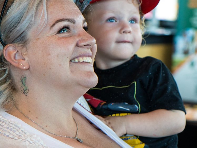 Image of A smiling woman with sunglasses on her head holds a young child wearing a red cap and black shirt. Both are looking up, appearing happy and engaged. The background is softly blurred indoors.