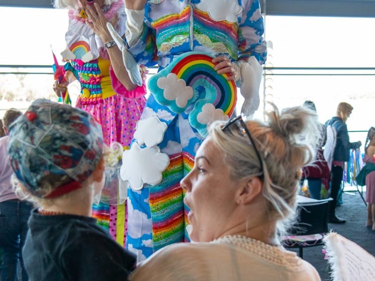 Image of A child and adult react with delight to two colourful performers dressed in whimsical costumes with rainbows, clouds, and large wigs at an indoor event with other people in the background.