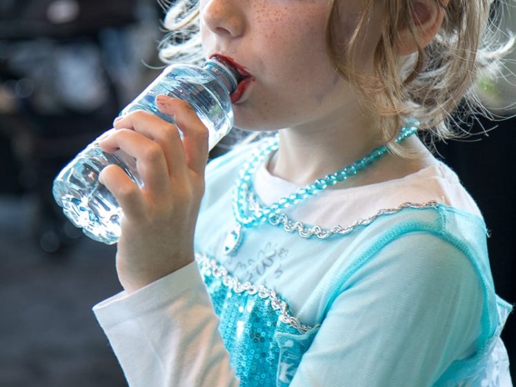 Image of A young girl dressed in an Elsa costume from the film Frozen drinks from a water bottle. She wears a tiara, beaded necklace, and has light brown hair with loose curls.