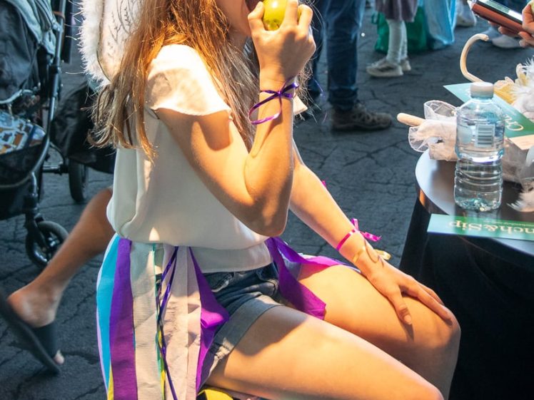Image of A girl wearing fairy wings, a headband with butterflies, and colourful ribbons sits on a stool and eats a green apple at a crowded indoor event.