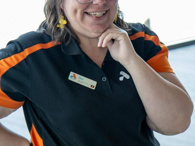 Image of A smiling woman with glasses and brown hair wears a black and orange collared shirt, a name badge, yellow earrings, and a yellow tutu. She is standing with one hand on her hip and the other touching her chin.