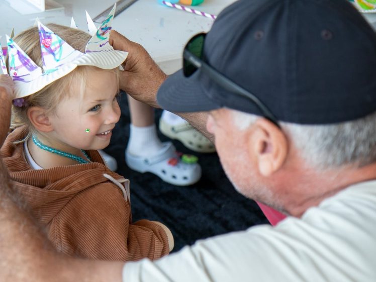 Image of An older man helps a young child wearing a handmade paper crown and a brown hoodie. The child, smiling and with a sticker on their cheek, looks up at the man during a craft activity. Other children are visible in the background.