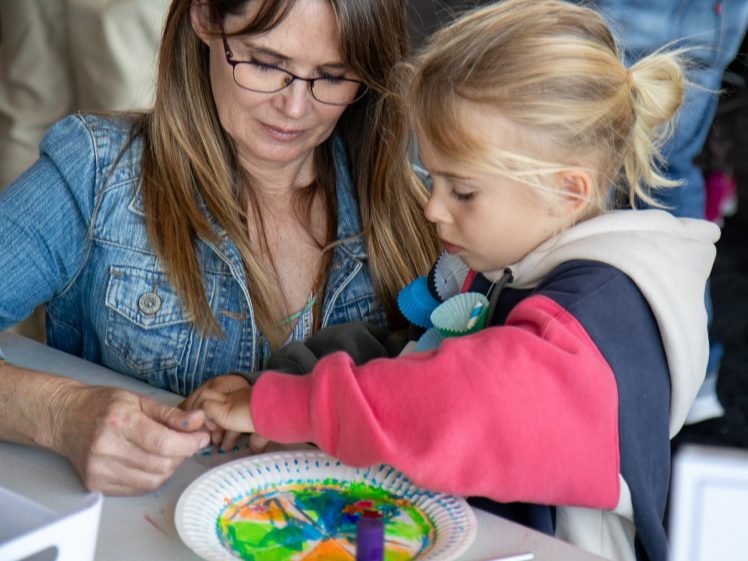 Image of A woman helps a young child create colourful art on a paper plate. The child is focused on the activity, and both are seated at a table with art supplies.