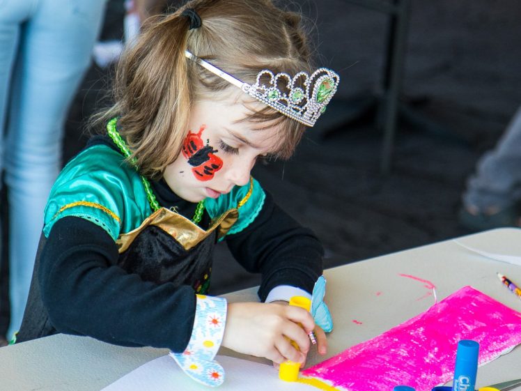 Image of A young girl wearing a tiara and fancy dress uses a glue stick on pink paper at a table. She has a butterfly painted on her cheek and appears focused on her craft activity.